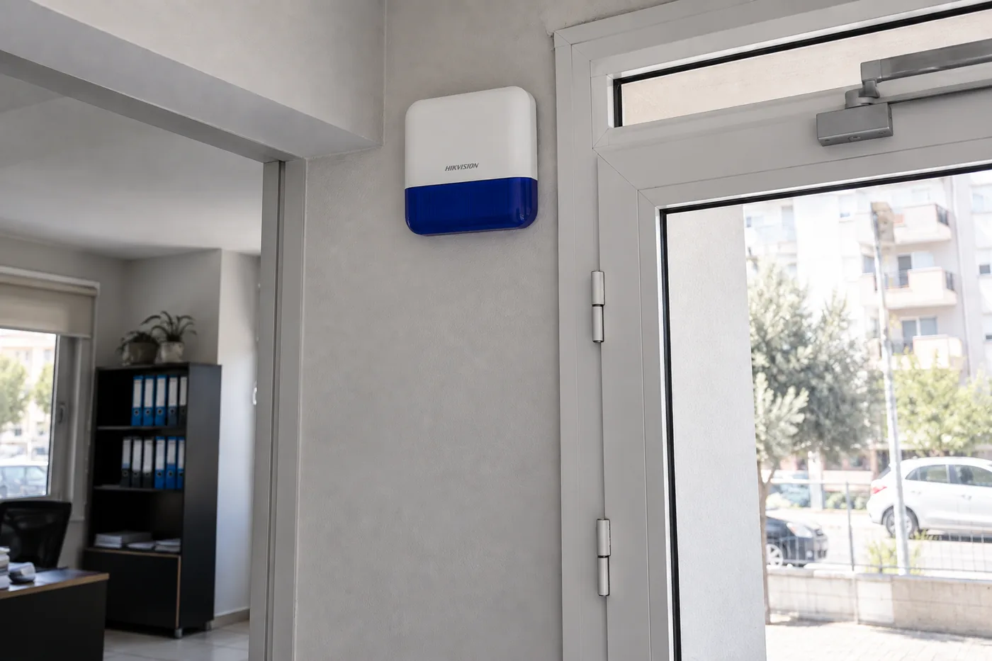 Küçük bir ofisin girişinde duvarın üst bölümüne monte edilmiş beyaz ve mavi Hikvision siren