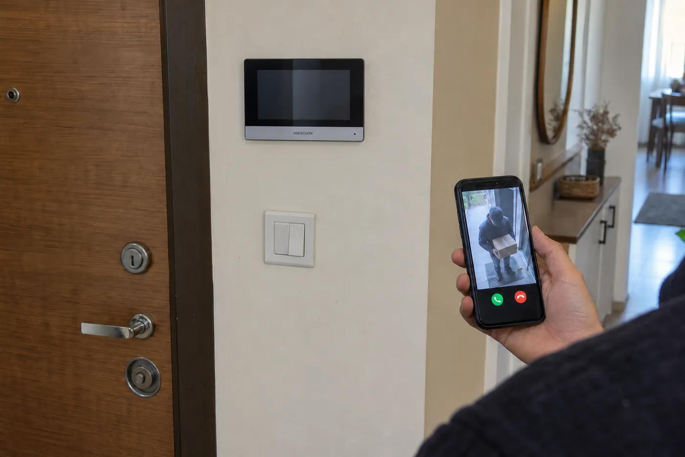 Daire antresinde telefonundan bina girişindeki kuryeyle görüşen sakin ve arka planda Hikvision DS-KH6320-WTE1 monitör