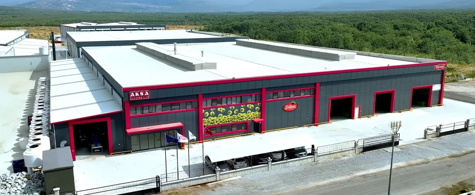 Aksa Zeytincilik'in Akhisar Süleymanlı OSB'deki üretim tesisinin havadan görünümü: antrasit cepheli, kırmızı şeritli fabrika binası; cephede AKSA Zeytincilik, Zeliva ve Yörem tabelaları; arkada zeytinlikler (firma görseli)